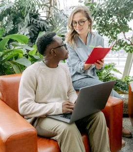Two professionals talking in a plant-filled office lounge, one with a laptop