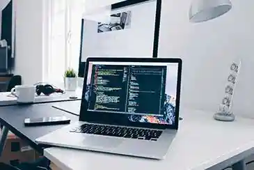 Laptop on a white desk displaying code in a dark editor, with a phone, headphones, and coffee mug nearby