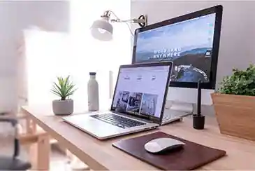 Laptop and monitor on a wooden desk showing a website and a Work From Anywhere wallpaper, with plants nearby