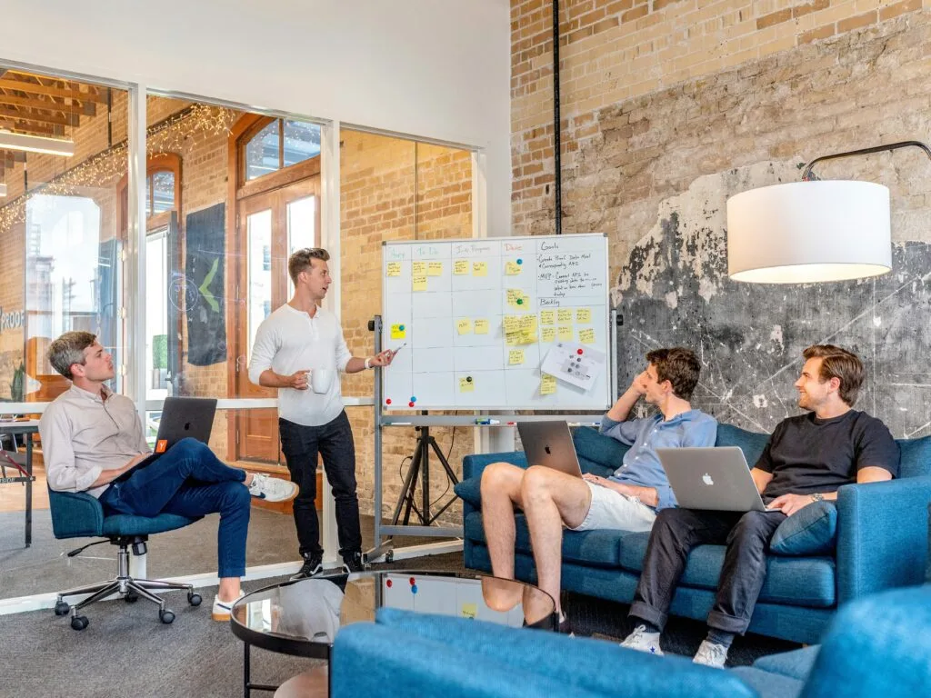 Four professionals planning at a whiteboard covered with sticky notes while three work on laptops